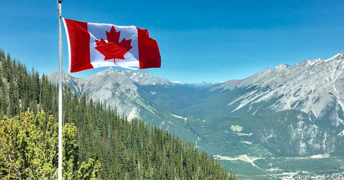 Canadian flag waving atop a scenic mountain view with blue skies and lush greenery.